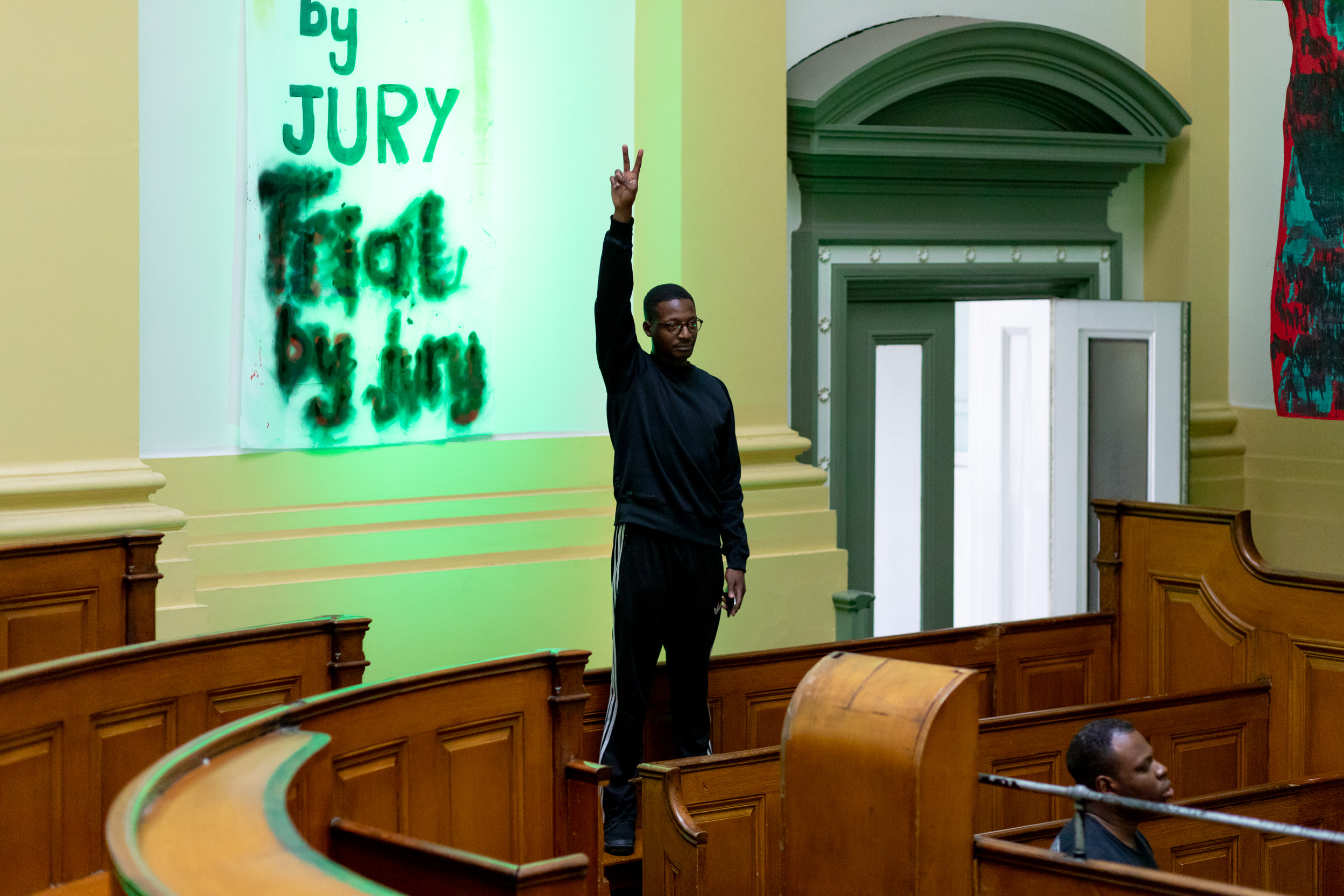 A black person wearing glasses, a black jumper and tracksuit bottoms stands with one arm in the air, their hand forms the peace sign. They are stood on a courtroom bench. The lighting is green. Behind them a fabric banner painted with green text reads 'Trial by Jury'.