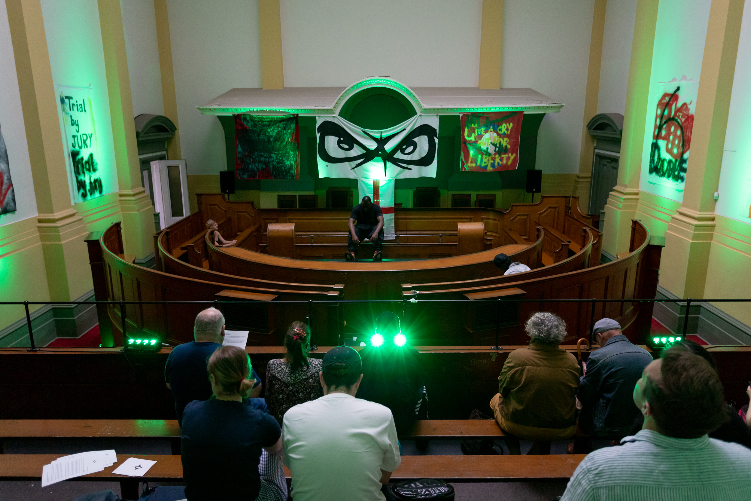 A courtroom space, with wooden seats and benches centred around a stage. A black person sits centre stage. Above and around them are fabric banners. The lighting throughout is green. Audience members sit on benches, watching and listening.