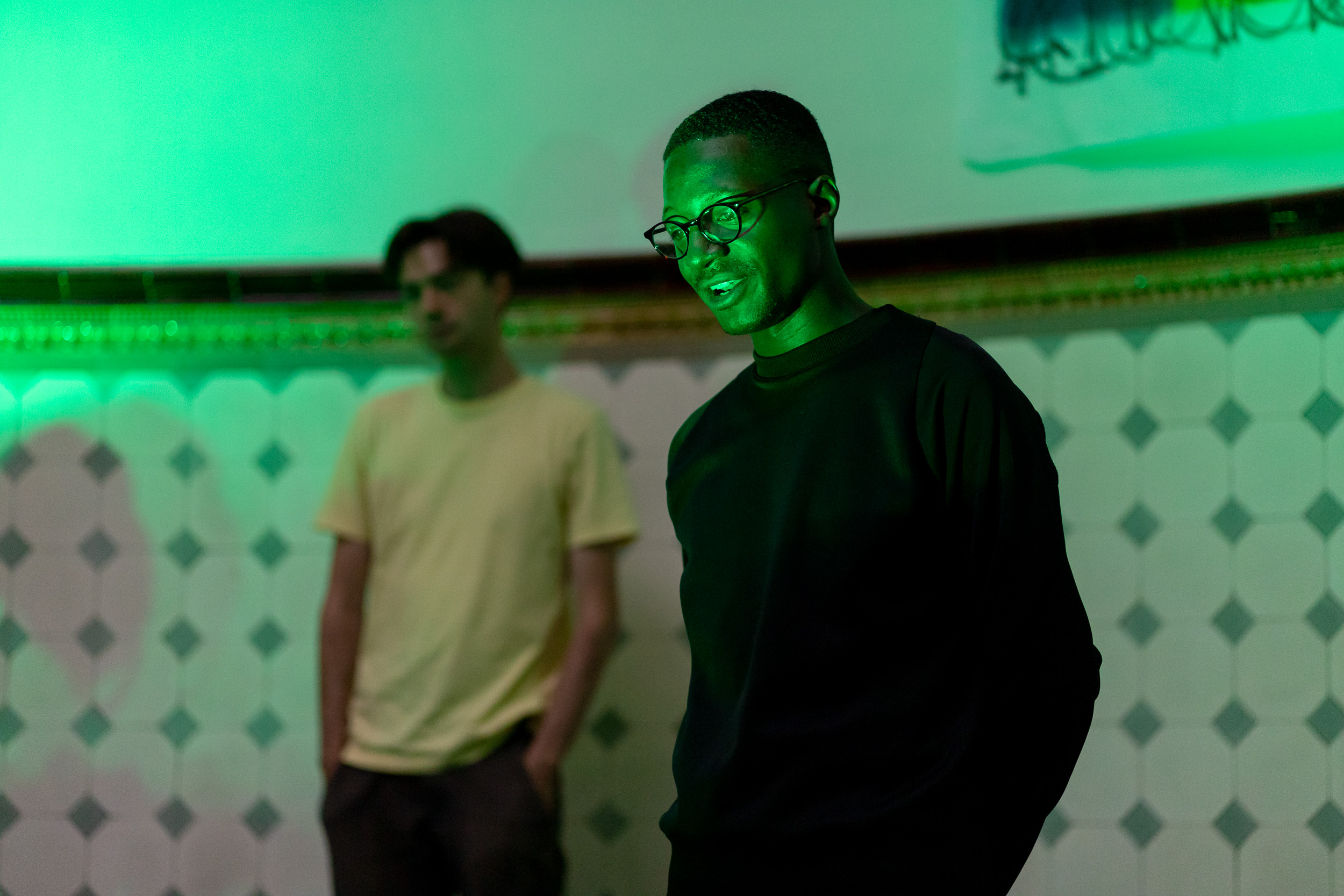 A black person wearing glasses and a black jumper is pictured in a space lit with green light. In the background another person is standing in the space, which has tiled walls.