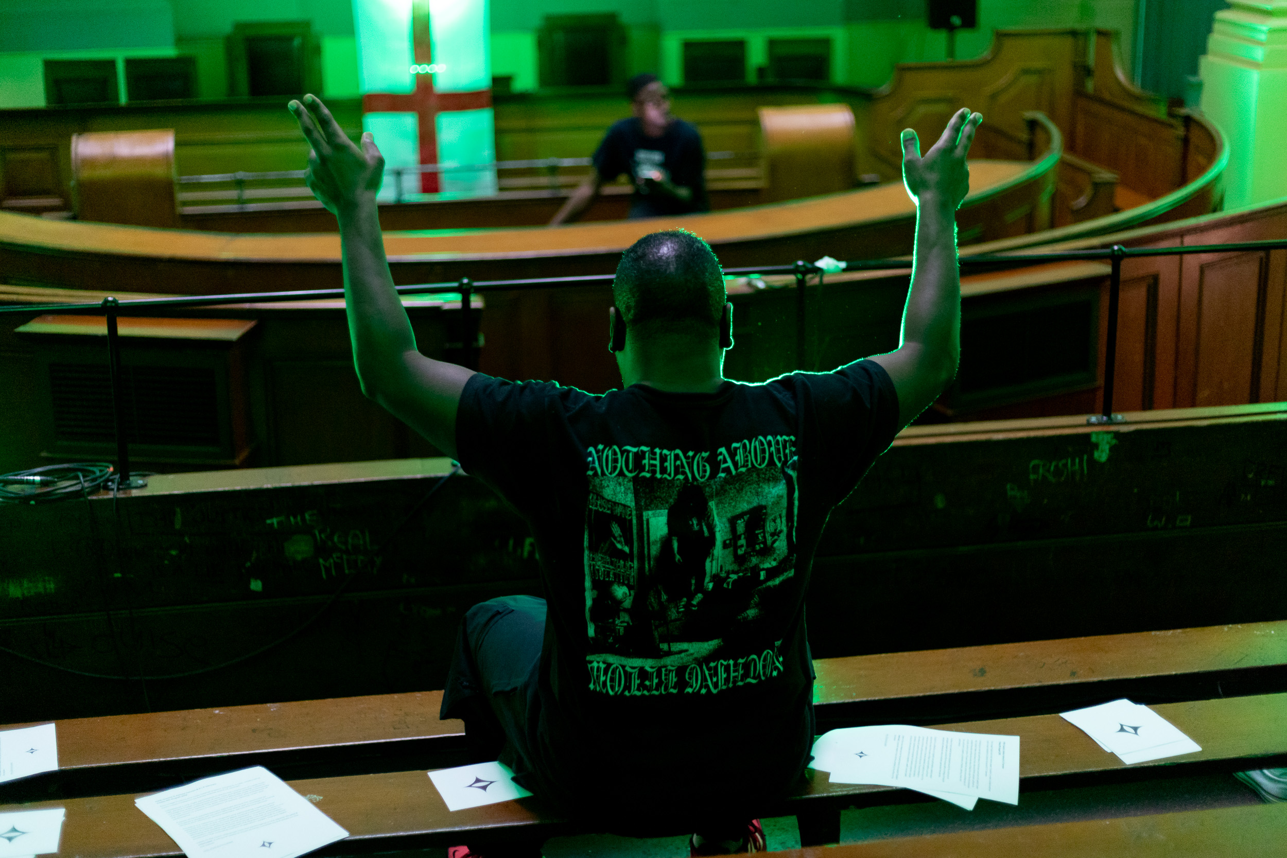 A person in a black tshirt stands with their arms raised up. Another person is seated in front of them. They appear to be in a concert hall or perhaps a place of worship.