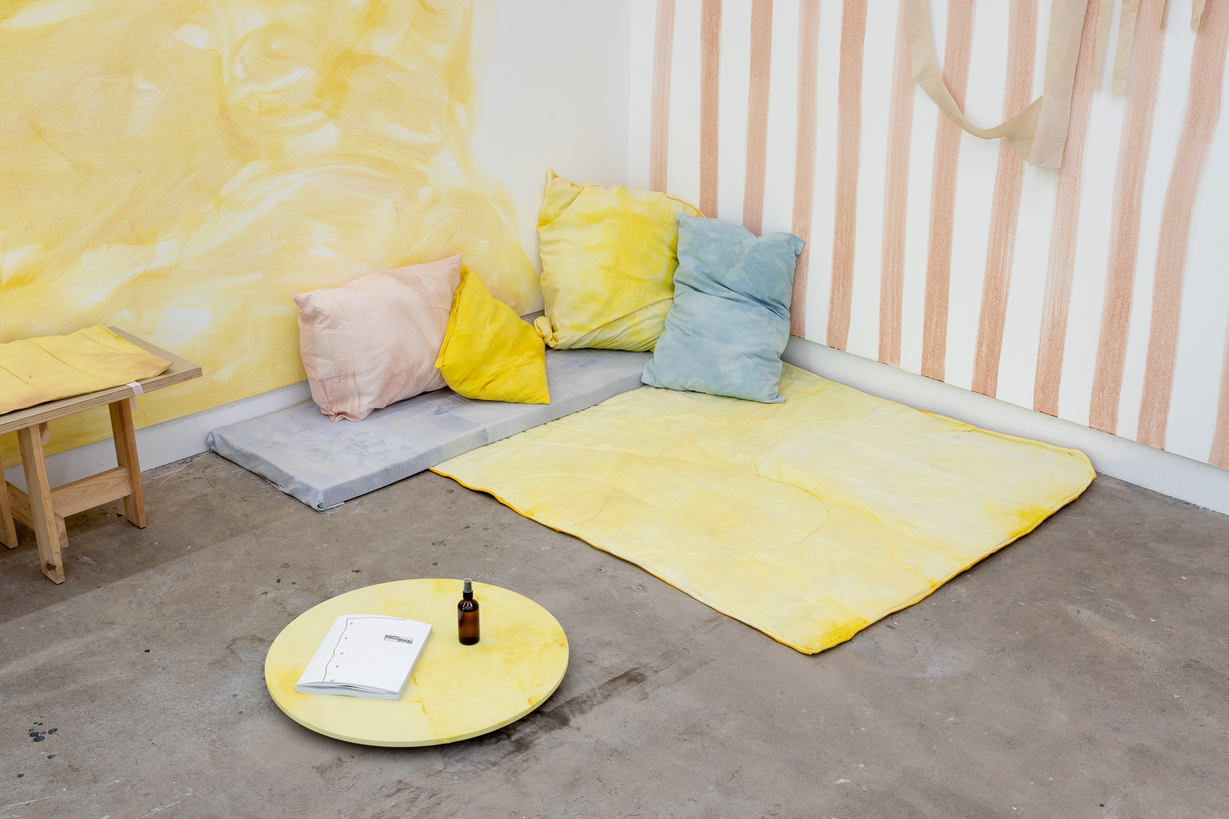 A corner of a gallery with cushions and mats to sit. A small wooden circle provides a table space. The walls are painted with swirls of yellow and pale pink stripes.