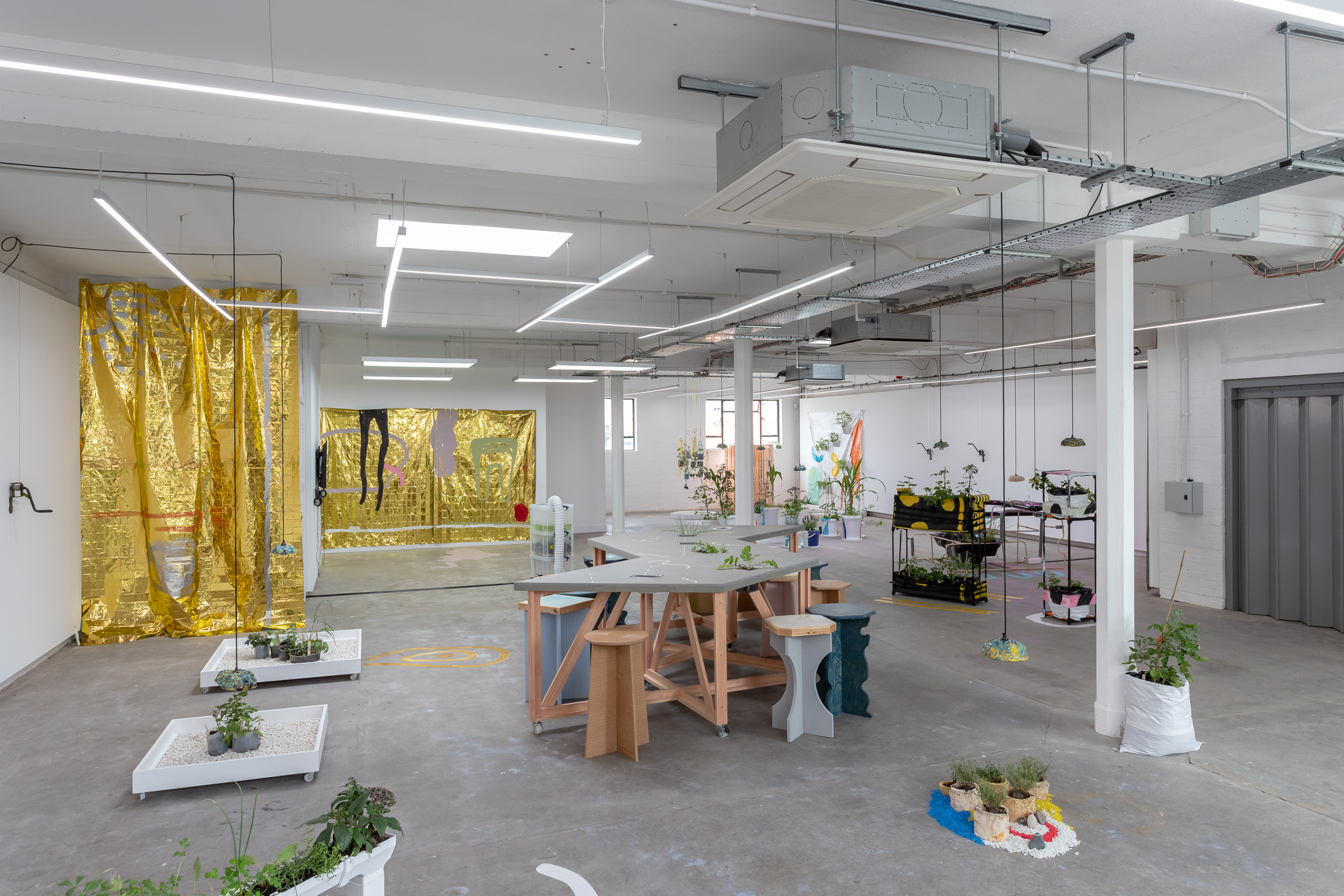 A view of a gallery with lots of different sculpture and artworks. Centrally there is a table and stools set up. There are several gold fabric wall hangings and curtains. Plants feature throughout the space.
