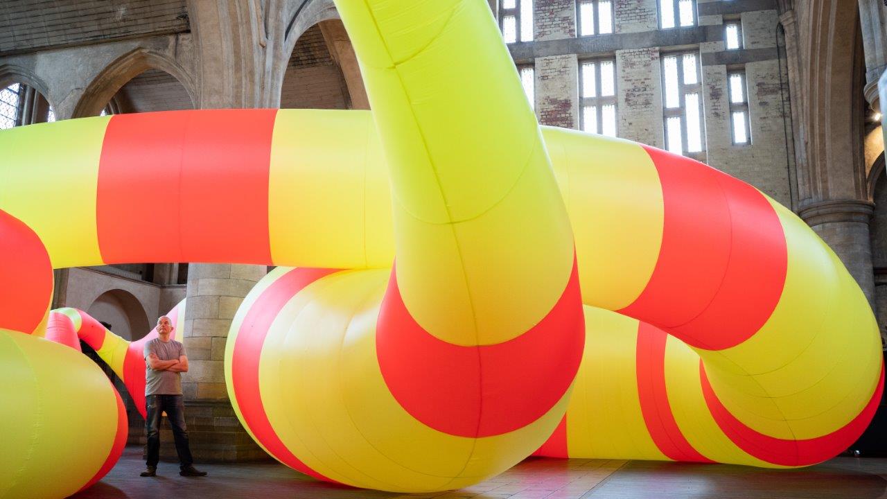 A white man stands looking up at a large inflatable artwork in yellow and red. It is snaking through a historic building, details of which are visible at the top and edge of the image.