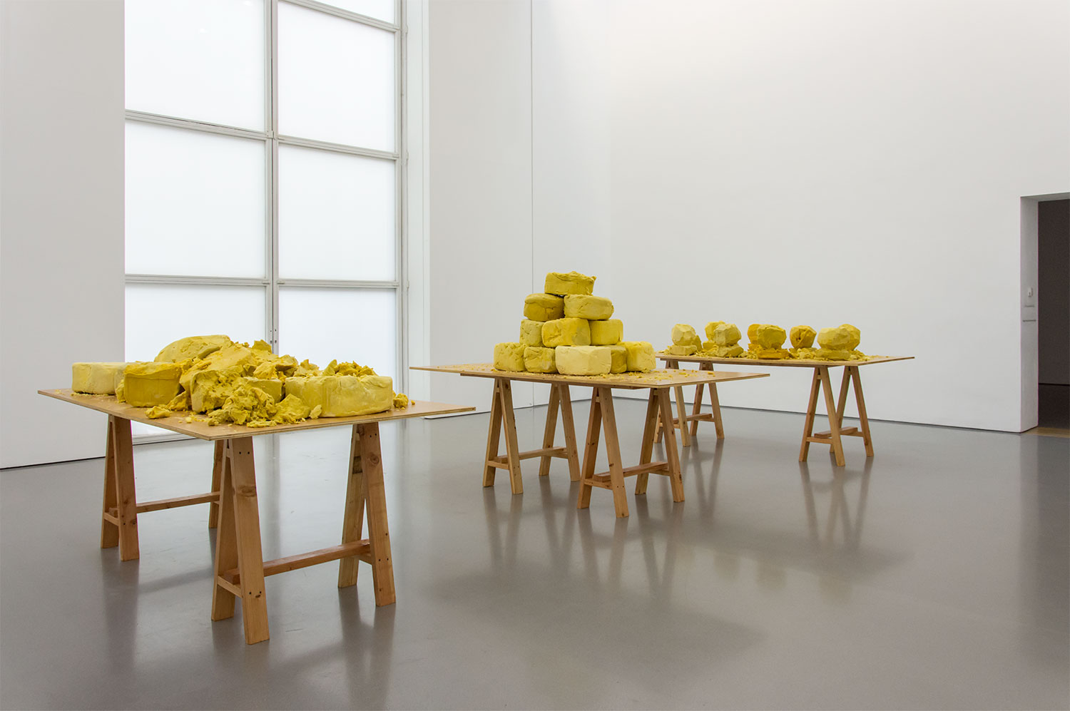 A gallery space with white walls and grey floor. In the gallery are three wooden tables which are all piled with lumps of shea butter in shades of golden yellow.
