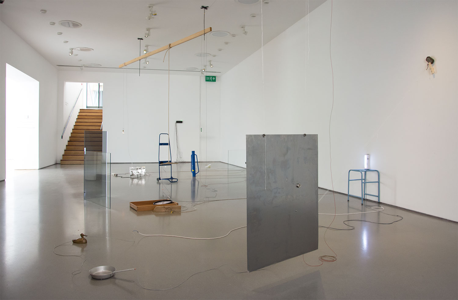 A gallery space with white walls and grey floor. Sculptures are positioned throughout the gallery, made up of sheet metal, glass and found objects. Cables trail throughout the space.