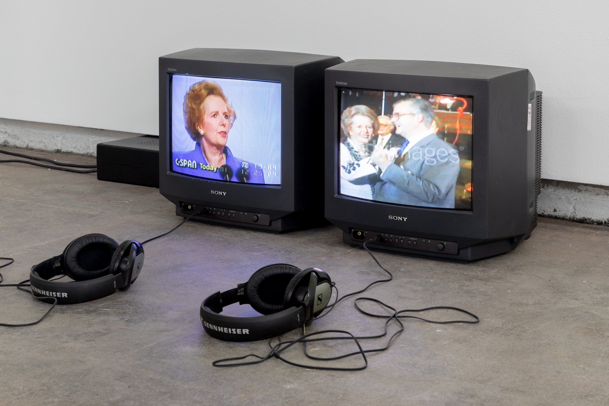 Two small cube TVs rest on the floor, with headphones plugged in. Both are playing films by the artist, on screen are two images of Maggie Thatcher.