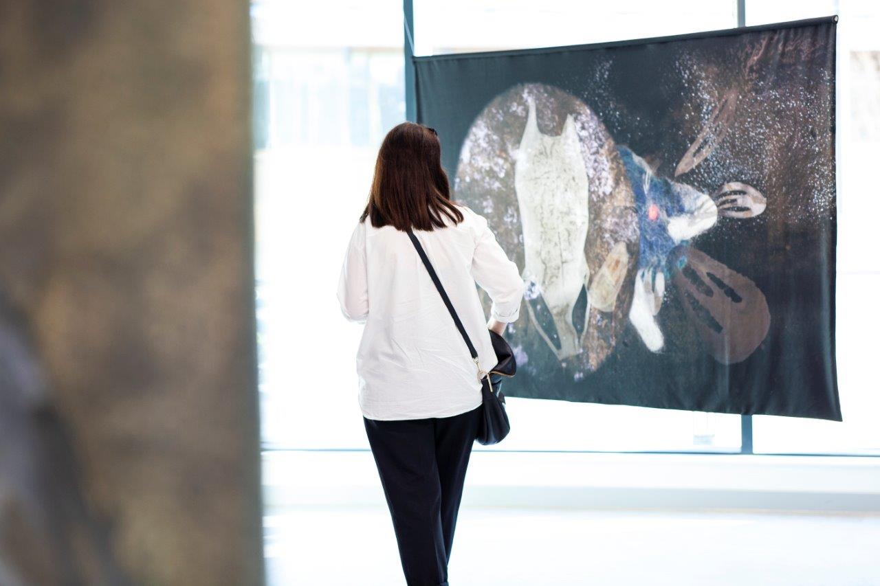 A person with brown hair wearing a white shirt stands in front of a painting in shades of black, purple and grey.