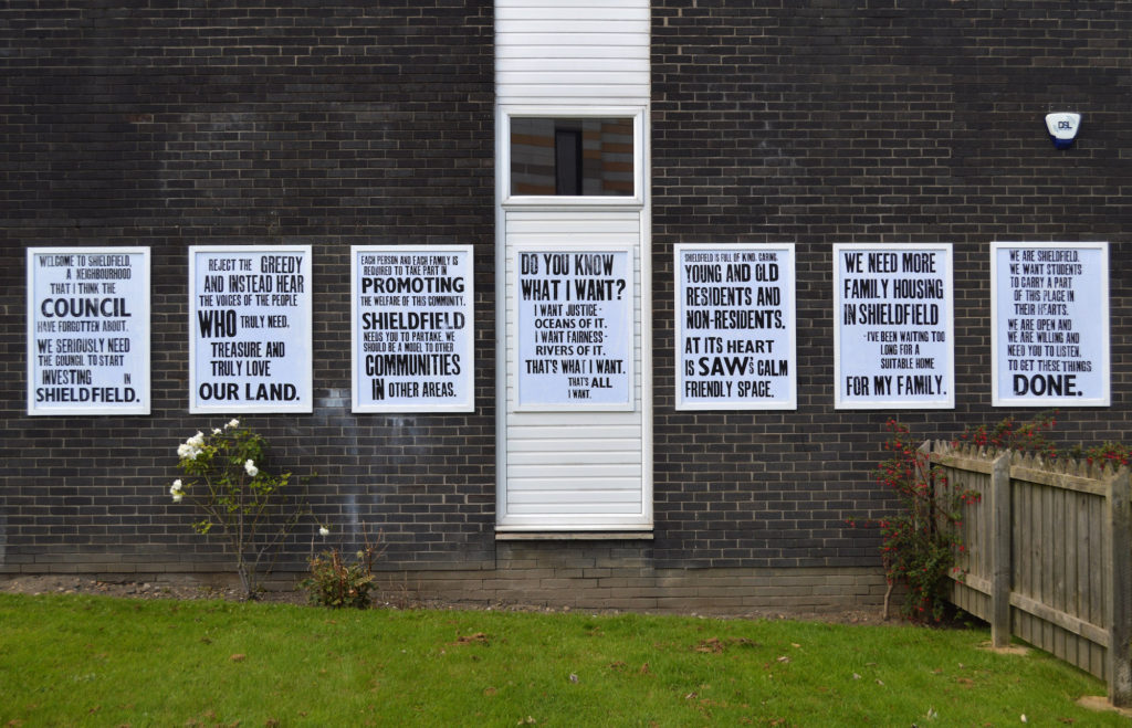 The outside brick wall of a 20th century building with a patch of white oanelling in the middle. On the wall and panel are displayed seven posters made up from words and slogans in different sied font. one phrase reads 'Treasure and truly love our land' another reads 'For my family'.