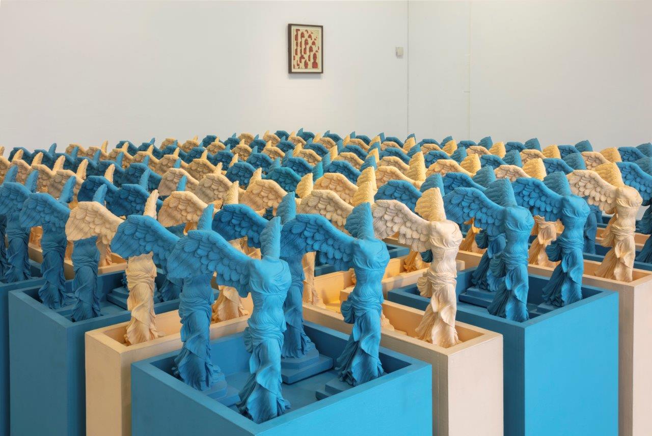 A series of sculptures in blue and yellow repeating a winged figure from classical sculpture.