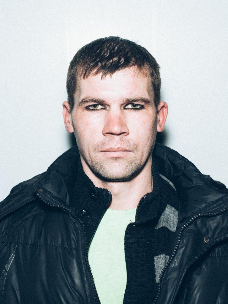 Portrait of a man looking directly into the camera. His hair is short, he wears a black coat with a pale green top underneath. His eyes are strikingly outlined by black eyeliner, emphasising their green-brown colour.