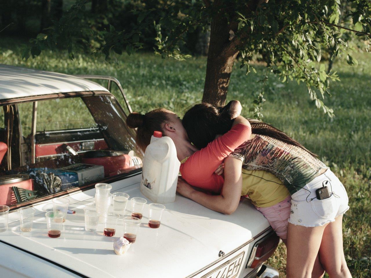 Two people embrace bending over the hood of a car. Balanced on the hood are cups and drinks.