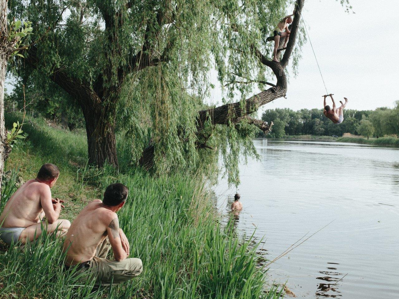 People relax at a riverside. Two people crouch, topless, in the long grass on the bank. Two others climb and swing from a tree over the river, while others are in the water.