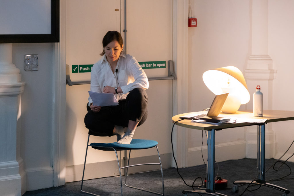 A person perches, crouched, on a blue chair. They are reading from a page, amplified through a clip-on microphone. Next to them is a laptop, lamp and water bottle on a table.