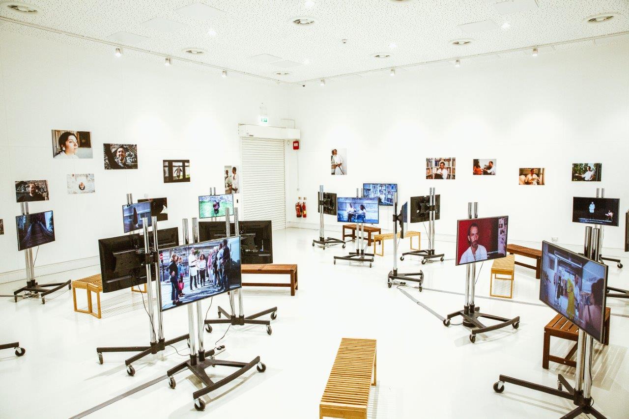 A gallery room filled with TV screens on metal legs and wheels. All are screening different videos. Benches are provided to sit. 