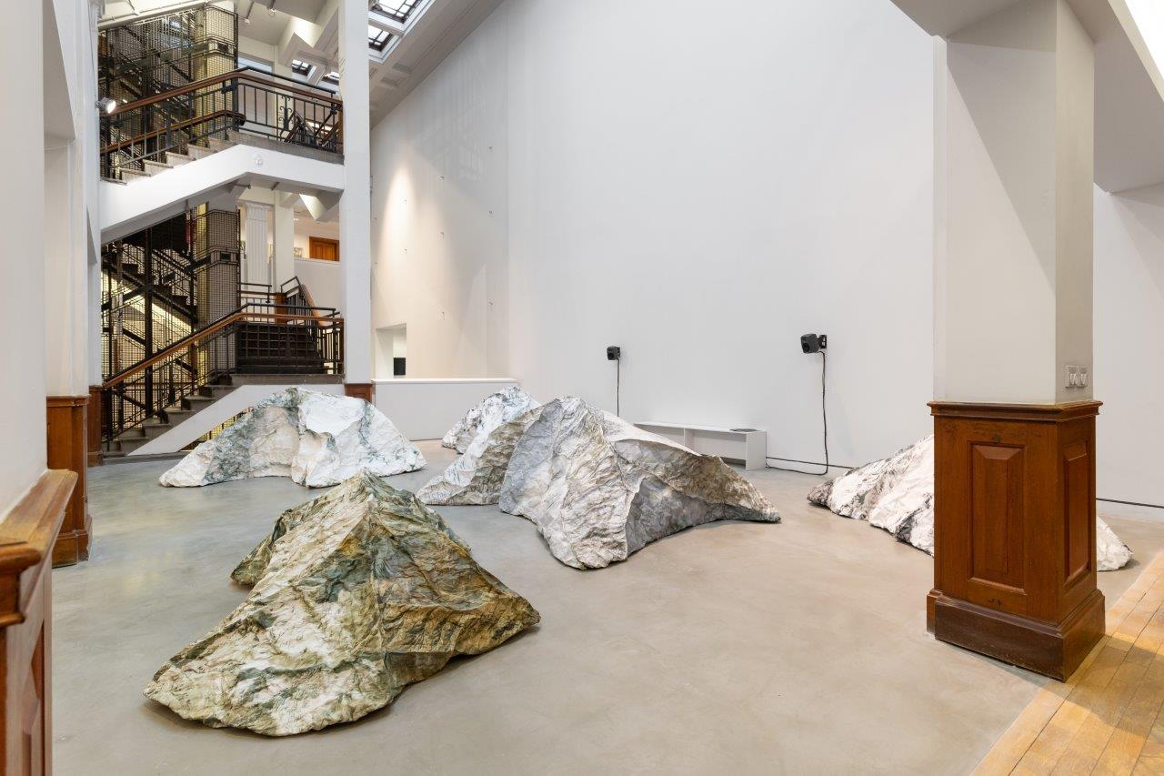Several sculptures in the atrium space of a gallery, the walls are white and the floor is grey. The sculptures look like mountains, in grey and gold.