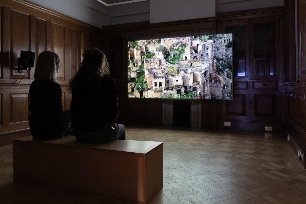 Two silhouetted figures sit to watch a video. The still on screen shows a ruined city.