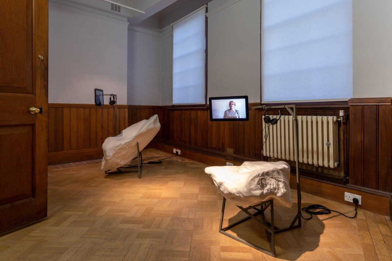 Two sculptures, with the appearance of stone or plaster, rest on metal legs. Between them a screen shows a video with a person on screen. Headphones are nearby to listen to the sound.