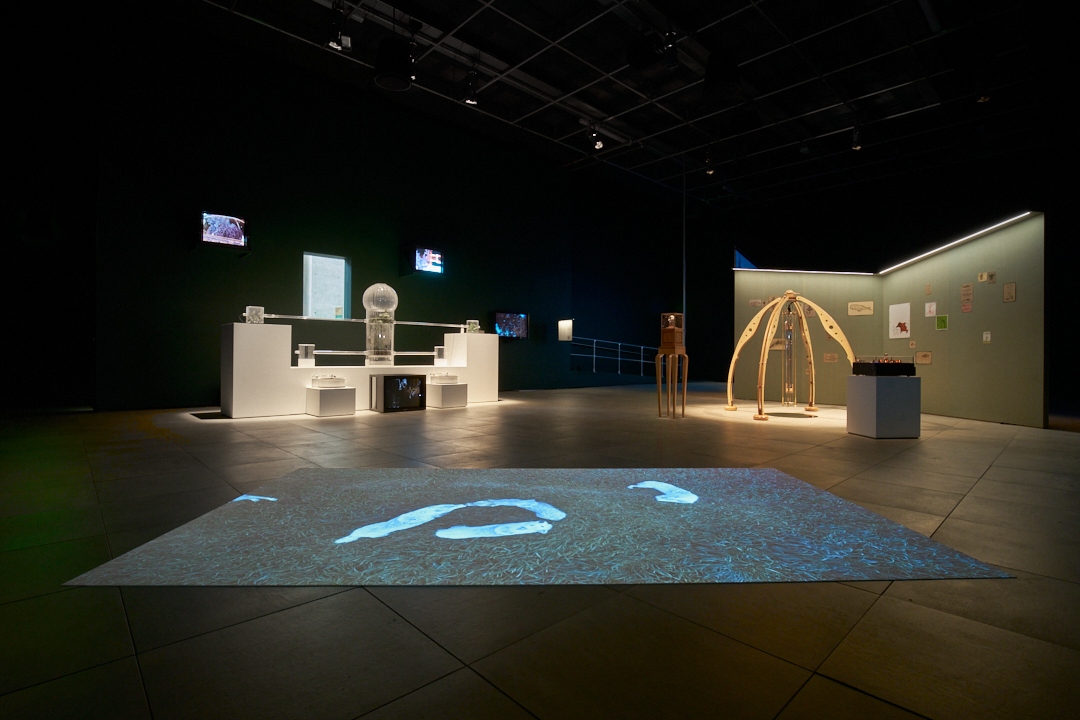 A large, dark space with two display areas to the left and right and a large image projected onto the floor in the foreground. 