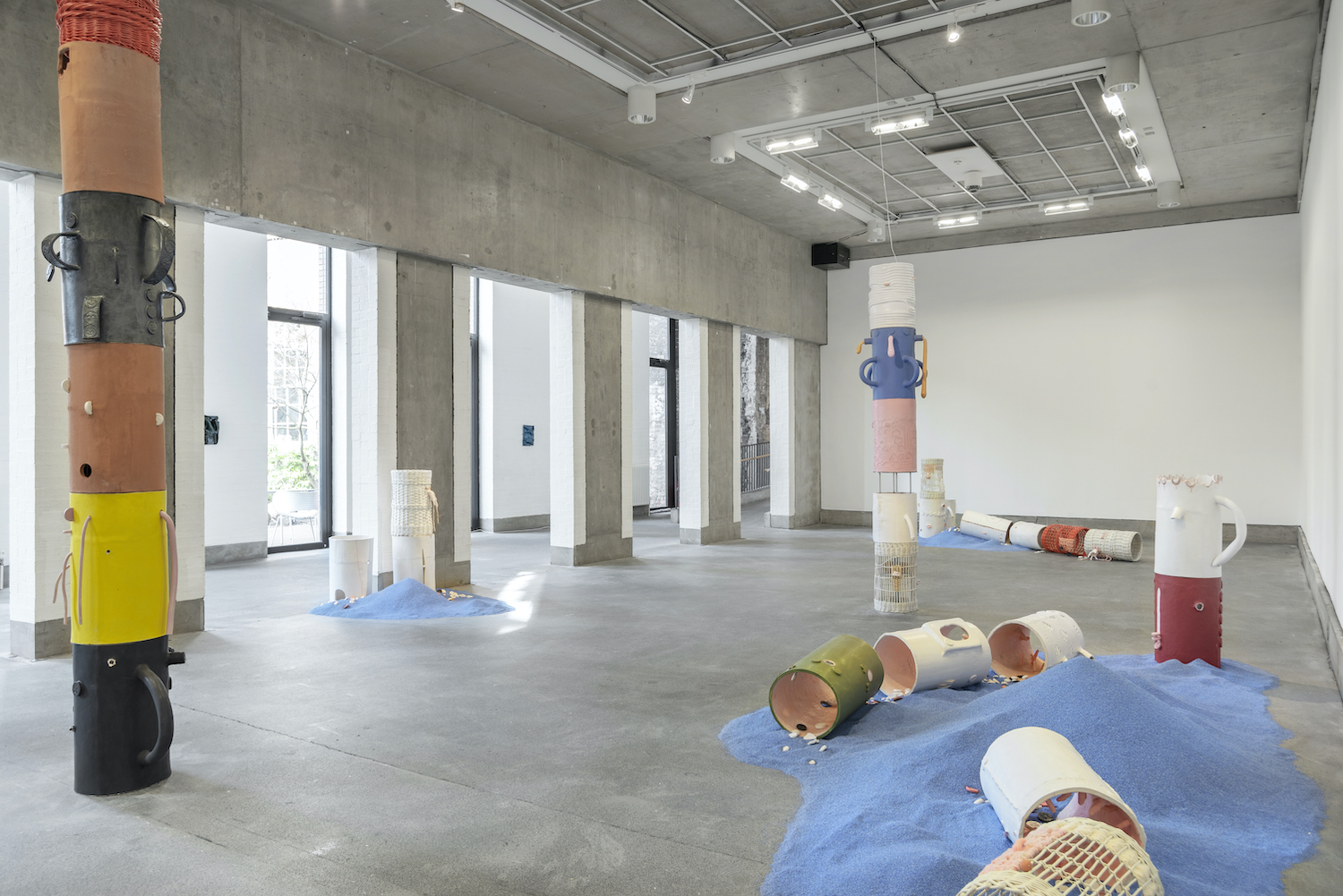 Multicoloured hollow cylinders are arranged in the gallery, in two columns to the left and right, and in the foreground arranged separately on the floor on a pile of blue sand.
