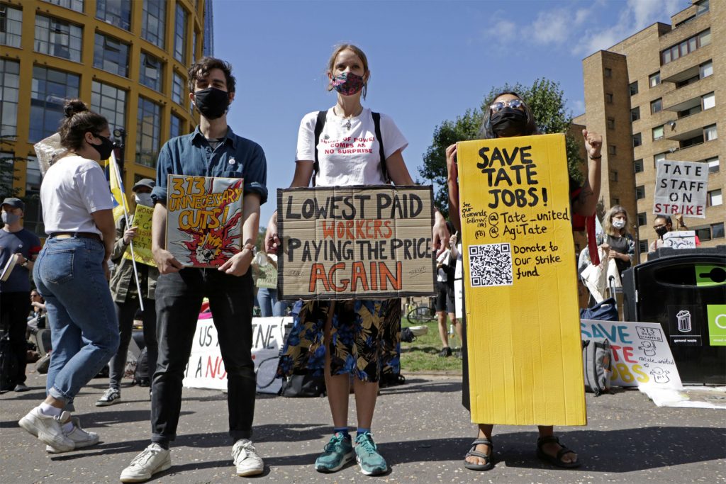 An image of a picket line outside Tate. Several members of staff stand together with signs and placards about low pay and saving jobs.