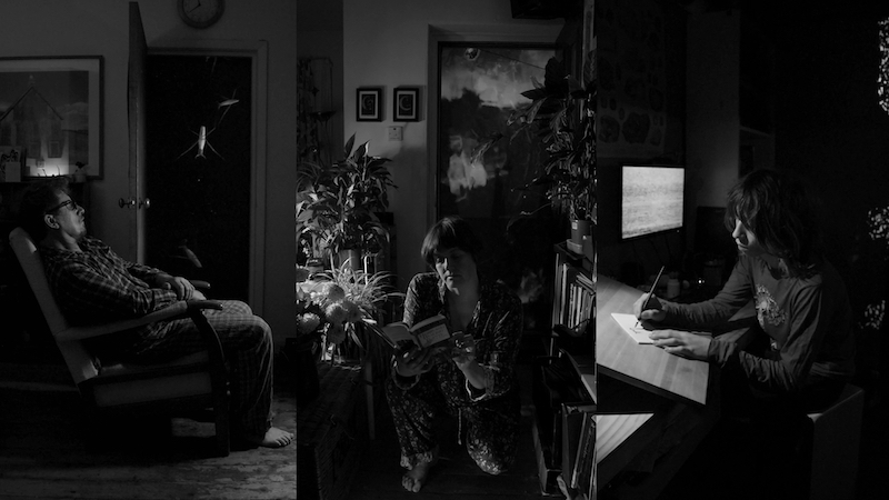 A screen is split into three. The image is black and white. On the left a middle aged man in pyjamas sits relaxed in a semi reclined pose in an arm chair. His arms are crossed in his lap and he has barefeet. Behind him a screen in a doorway has images of pondskaters on it. The centre image shows a middle aged white woman wearing pyjamas kneeling down between a table and a bookcase. There are flowers on the table. She is reading a book. On a screen behind her clouds can be seen. The right image shows a young white boy sat drawing on paper next to a screen showing static pattern. Behind him is a projected image of foam on water.