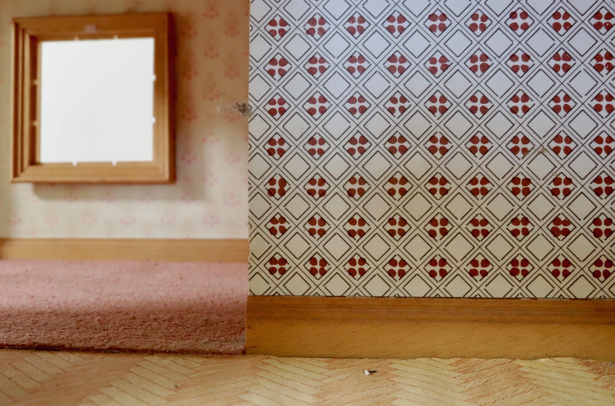 A woman holds a vanity mirror to obscure her face, The reflection in the mirror shows the doors of a dolls house. The woman has a grey knitted head piece on which falls to her shoulders. She is wearing a stripey pyjama top Looking through a doorway in a dolls house with no door to the left of frame. To the right is a close up of the wallpaper with a red cross and square pattern. The room has a rose coloured rug and a square window with a wooden frame. A screen is split into three. The image is black and white. On the left a middle aged man in pyjamas sits relaxed in a semi reclined pose in an arm chair. His arms are crossed in his lap and he has barefeet. Behind him a screen in a doorway has images of pondskaters on it. The centre image shows a middle aged white woman wearing pyjamas kneeling down between a table and a bookcase. There are flowers on the table. She is reading a book. On a screen behind her clouds can be seen. The right image shows a young white boy sat drawing on paper next to a screen showing static pattern. Behind him is a projected image of foam on water. This image shows the view through the glass of french style doors. Inside the light cast an orange glow across the whole room. Directly through the doors on the opposite wall are two windows, closed with a sofa in from of them. There is a large cushion on the sofa. To the right on the wall is a picture of a woman face drawn. The ceiling is wood panelled and a light on a chain hangs from the centre. We see an image looking over the sea towards the mountains in the distance. Close up in the sky are two sea birds with black tips. They are flying separate although at the same height. The sea is calm with a very small swell, no waves. The sky has thin white clouds.