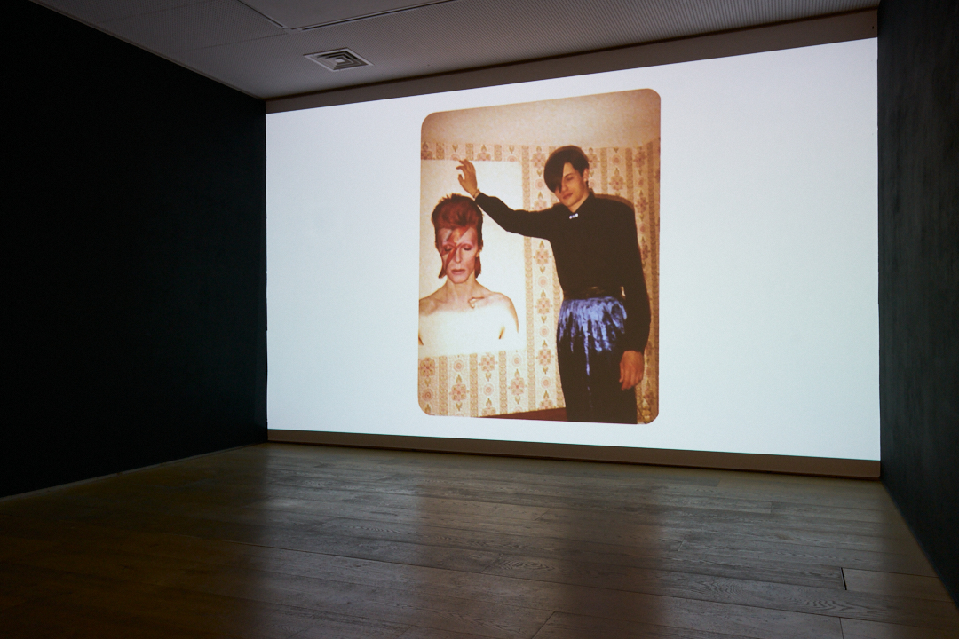 An old photograph projected large onto a white wall shows a young person in dark clothes posing beside a Ziggy Stardust poster.