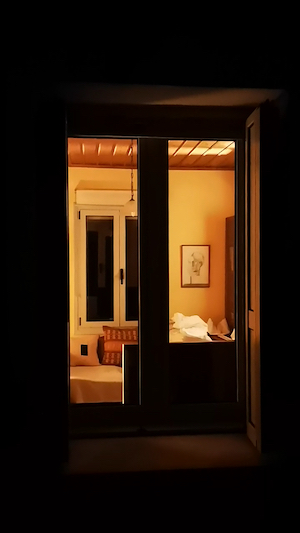 This image shows the view through the glass of french style doors. Inside the light cast an orange glow across the whole room. Directly through the doors on the opposite wall are two windows, closed with a sofa in from of them. There is a large cushion on the sofa. To the right on the wall is a picture of a woman face drawn. The ceiling is wood panelled and a light on a chain hangs from the centre.