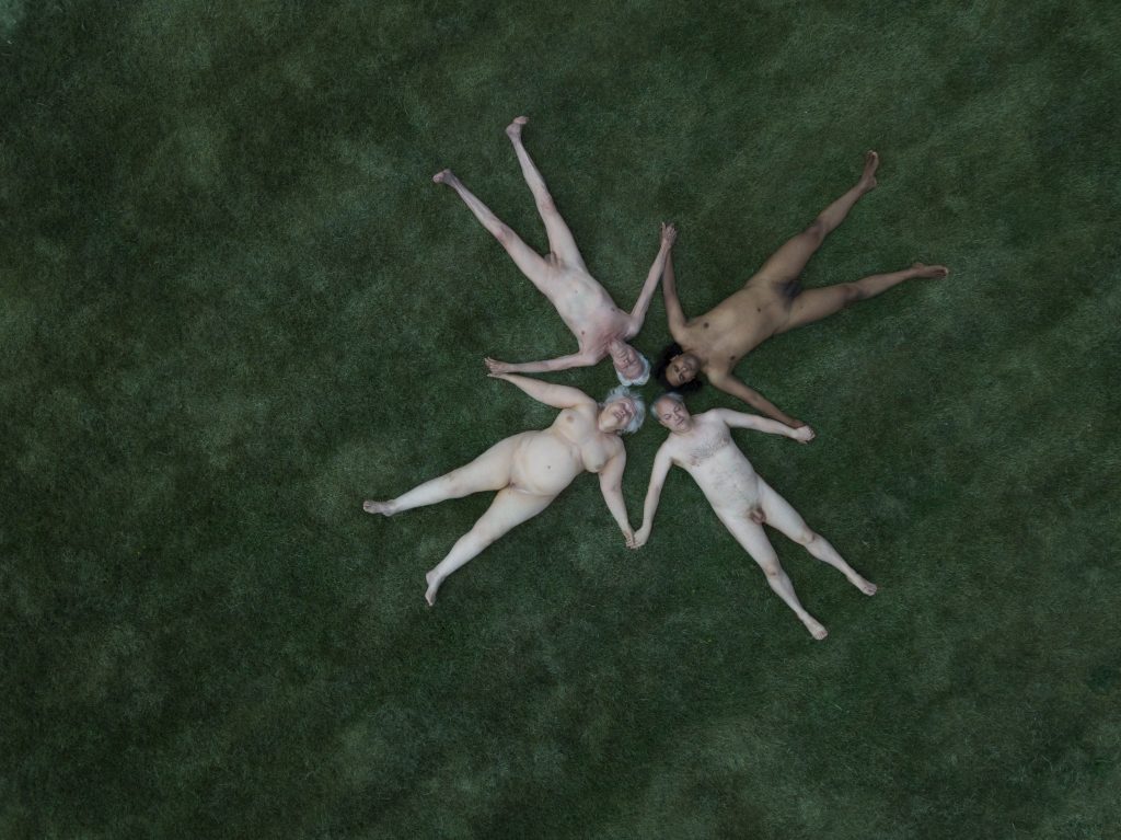 A photogarphic image of four nude people, lying on a green background. Their bodies are arranged in a star formation, seen from above.