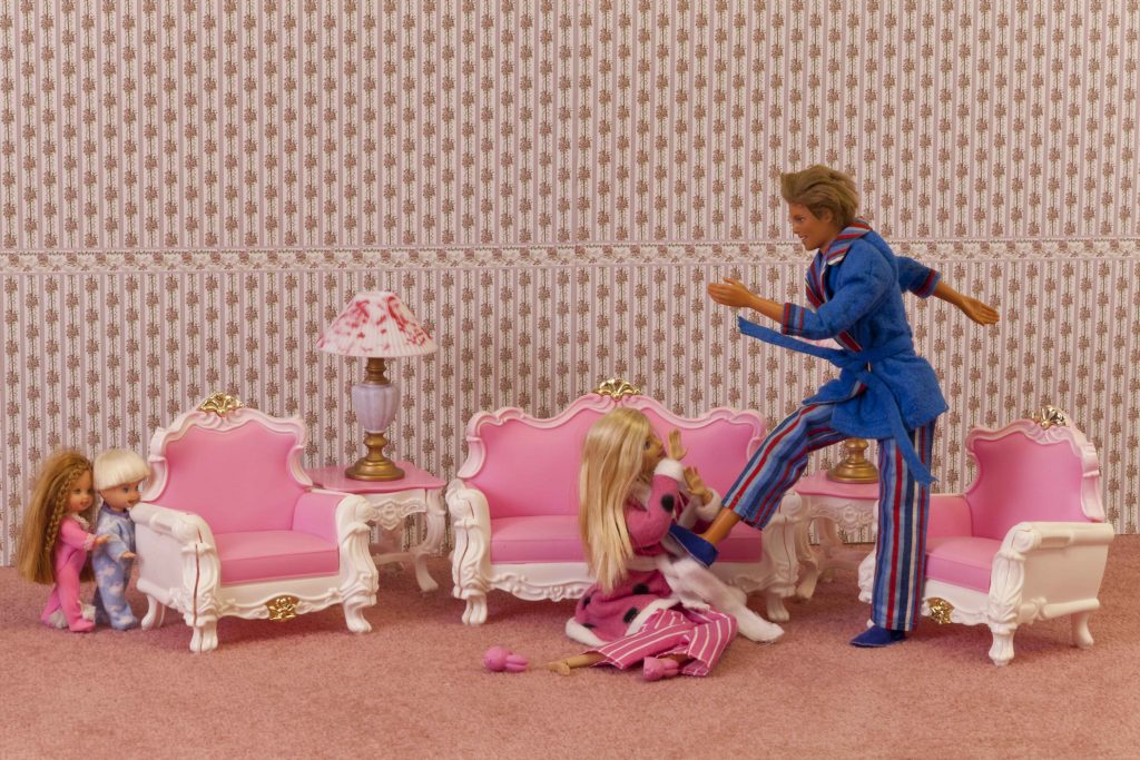 A scene composed with Barbie dolls, showing a Ken doll assaulting a barbie. Two child dolls watch the scene from behind a sofa.