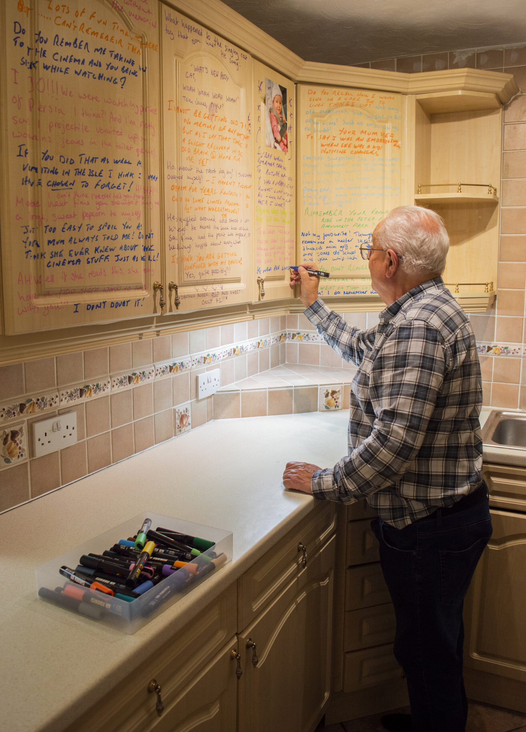 An older white man with white hair, glasses and wearing a checked shirt is facing away from the camera and writing on kitchen cabinet doors using paint markers. There is multi coloured writing all over the cabinets and a box of markers in the foreground.