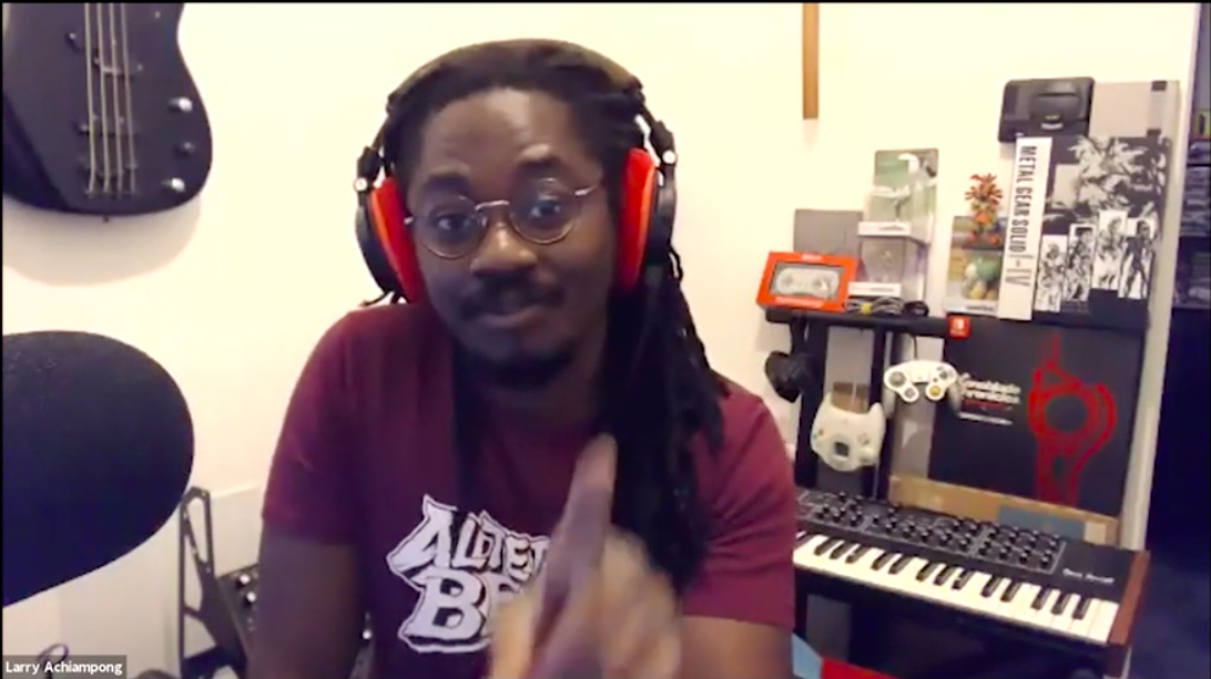 A Black man looks directly at the camera, his hand blurred at the bottom of the frame. He wears red over ear headphones and sits in front of an electric piano.