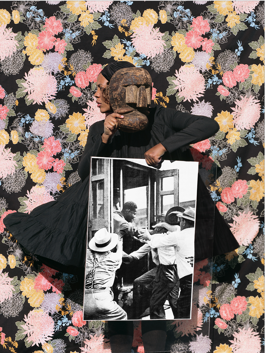 A black woman dressed entirely in black including a head covering holds a wooden mask near her head but her face can be seen to the left looking away. In her other hand is a large black and white photograph of men pulling another man out of a doorway. She stands in front of a background of black with multicoloured floral prints.
