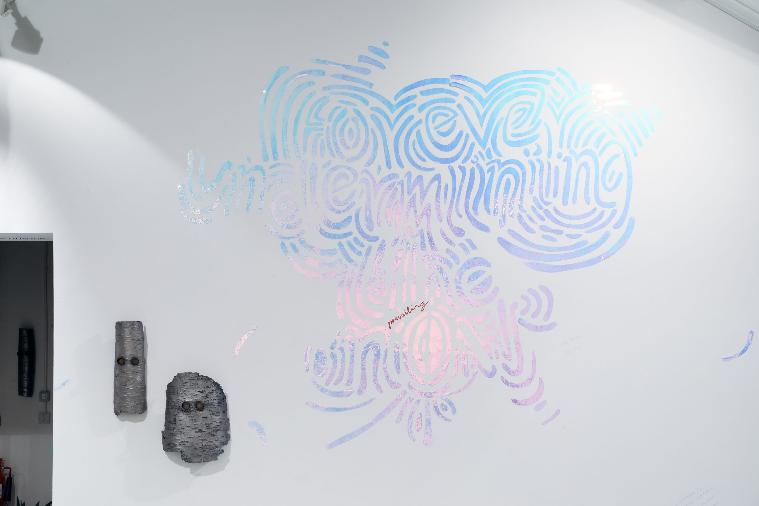 Installation wall painting with words 'Forever Underminging the prevailing Now' in blue and pink. Two metal mask sculptures wall mounted to the left.