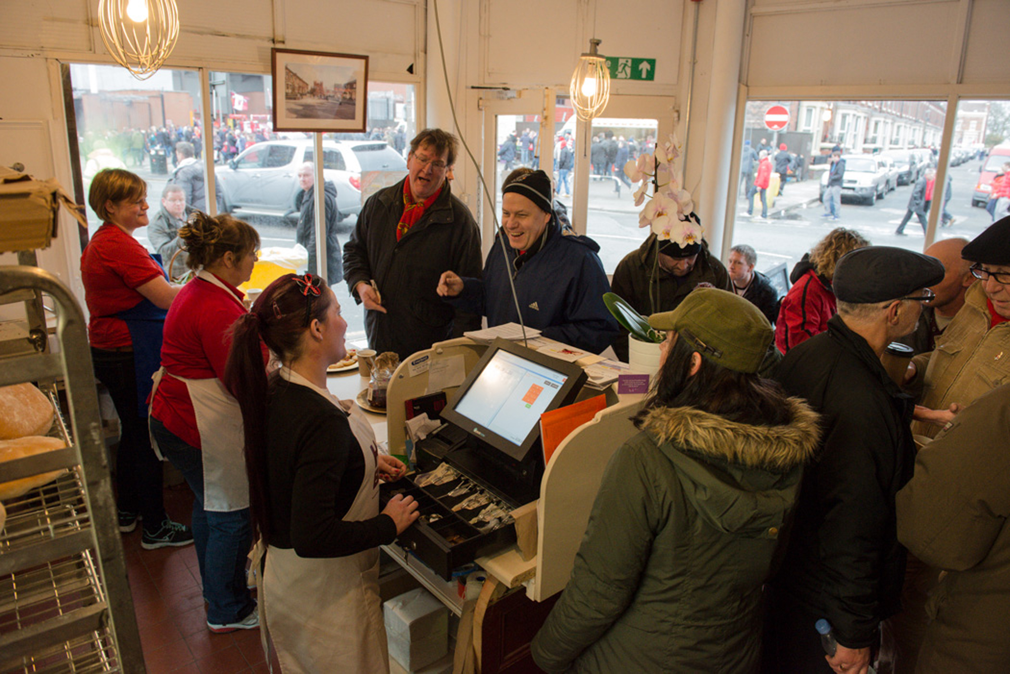 A group of people gathered inside a bakery. One person is at the till, behind them are metal shelves containing loaves of bread.