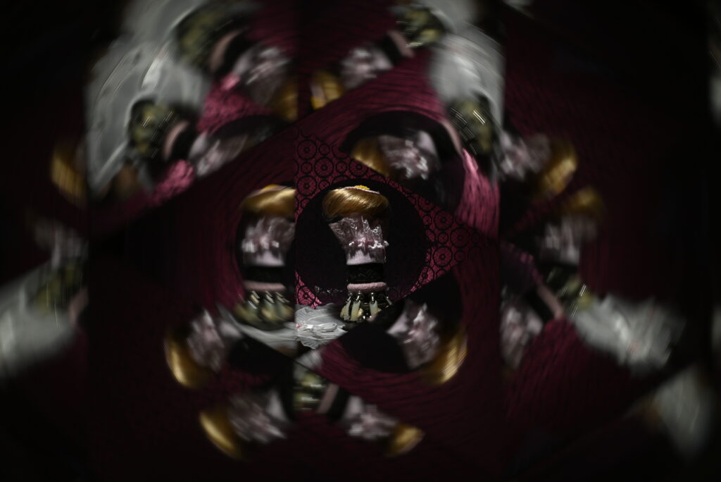 Set against a black background a kaleidoscope tesselates parts of an image. In the centre a small, clear, image of a mannequin complete with a wig, white cotton lace collar, and green tassels is visible.