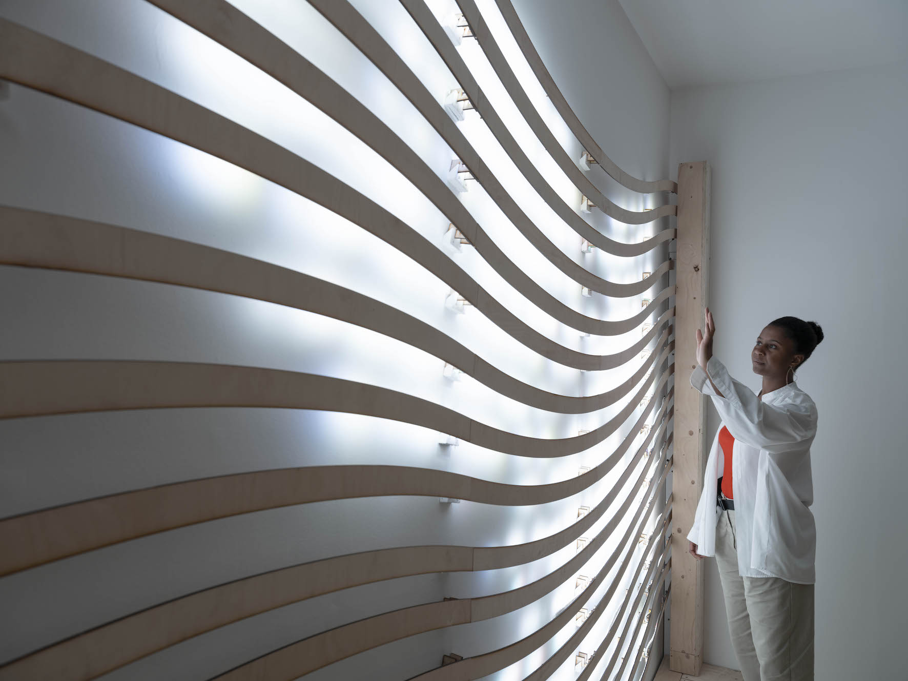 The artist stands beside a wavy sculptural wall installation, backlit.