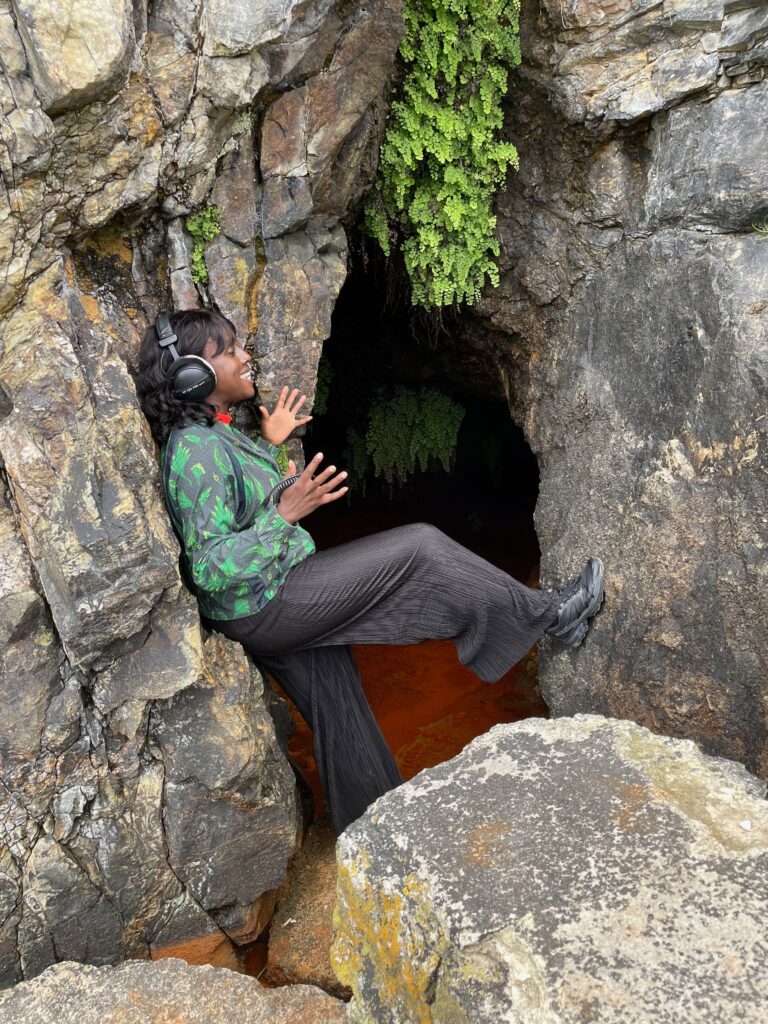 The artits stands with her foot up on a cave wall, with headphones on and appearing to speak or sing.