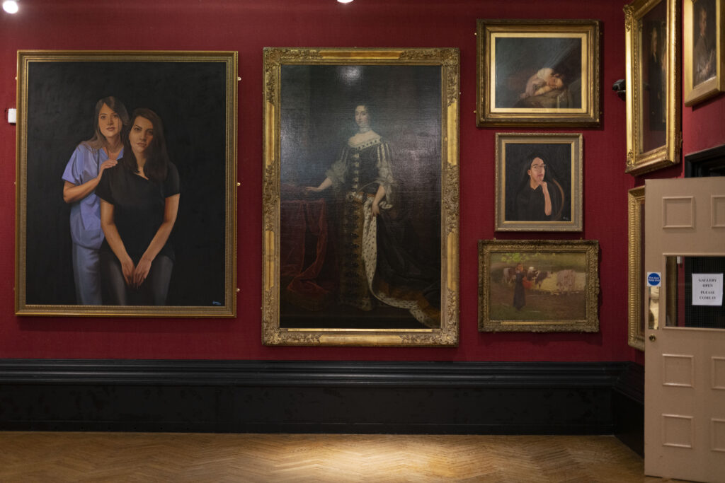 The image shows the portrait gallery at Blackburn Museum and Art Gallery, with classical paintings hung floor to ceiling in gilded frames against a red wall. Interspersed between the classical paintings are contemporary portraits of South Asian women.