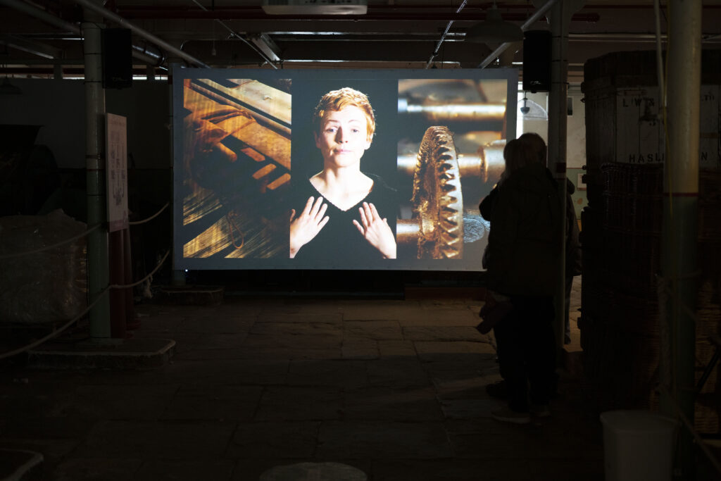 A screen in the middle of a room showing a three channel video can be seen, with the video showing parts of mechanical looms on either side of a figure pleading to the viewer.