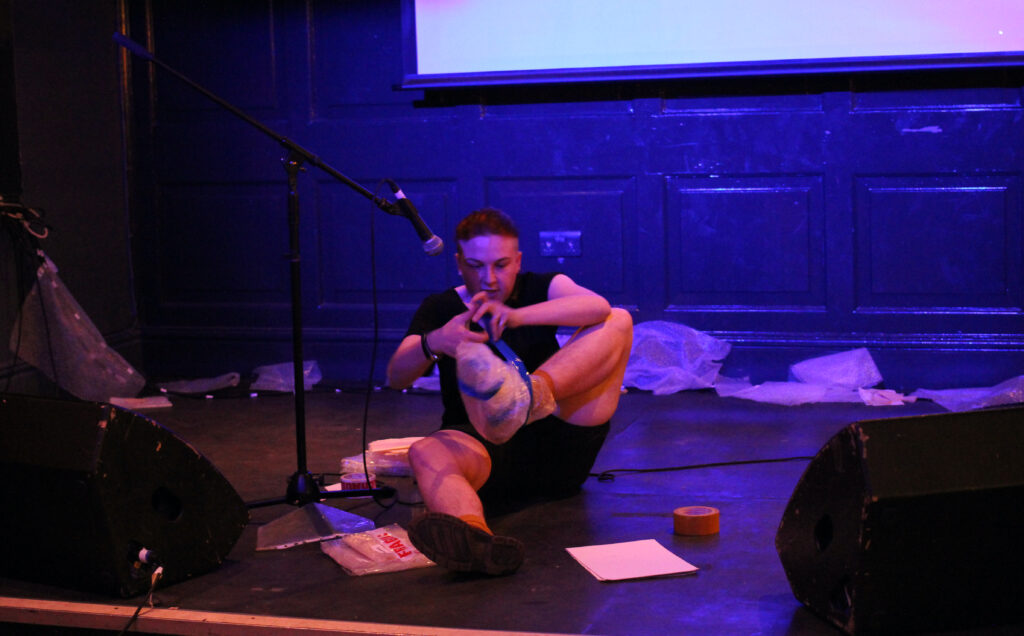 The artist sits on a stage with a microphone, taping bubble wrap around his left foot and ankle. 