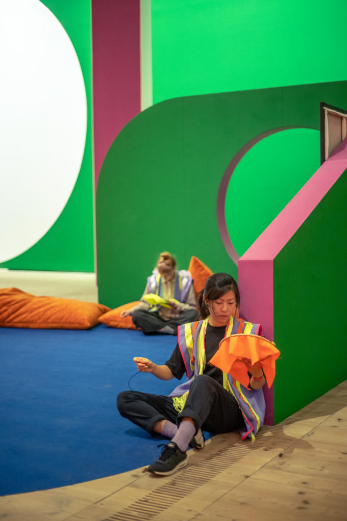A gallery space made up out of large, brightly coloured forms. Sitting on the floor and on cushions, sit women who are absorbed in a task of sewing.