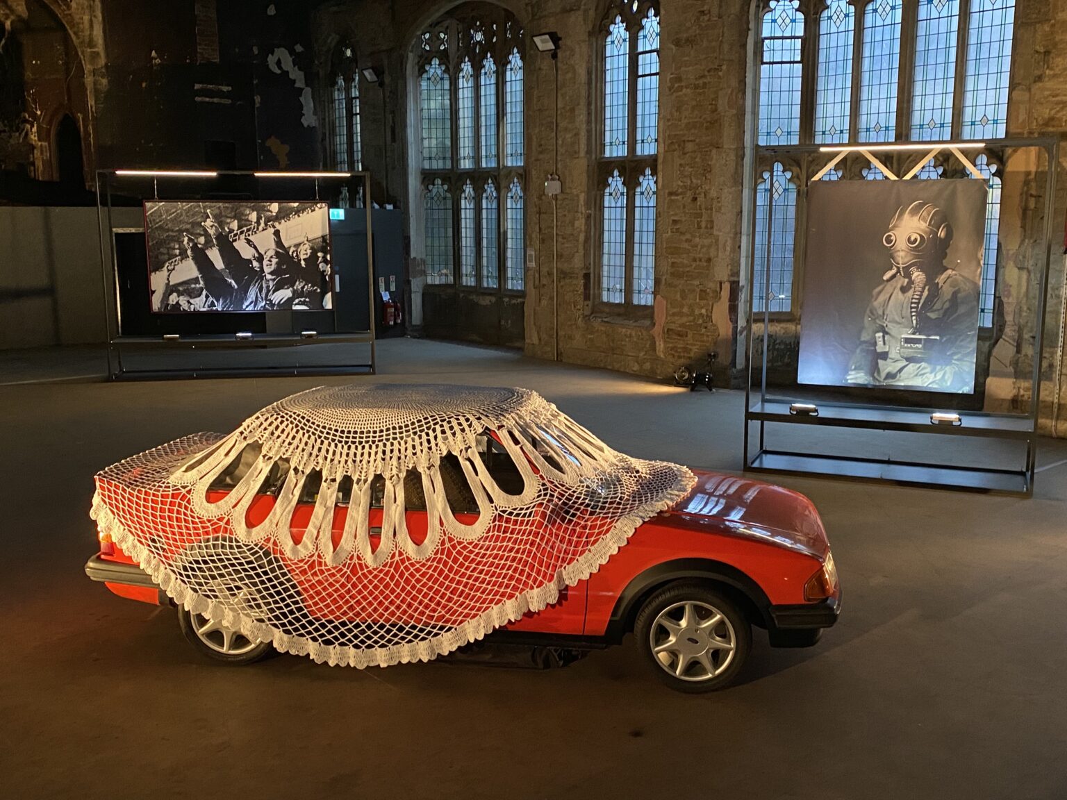 An image of a derelict, formerly grandiose space with a red car covered in a giant doily in the foreground, a photographic portrait of a man in a gask mask in the middle ground, and a tapestry of football fans chanting in the background.