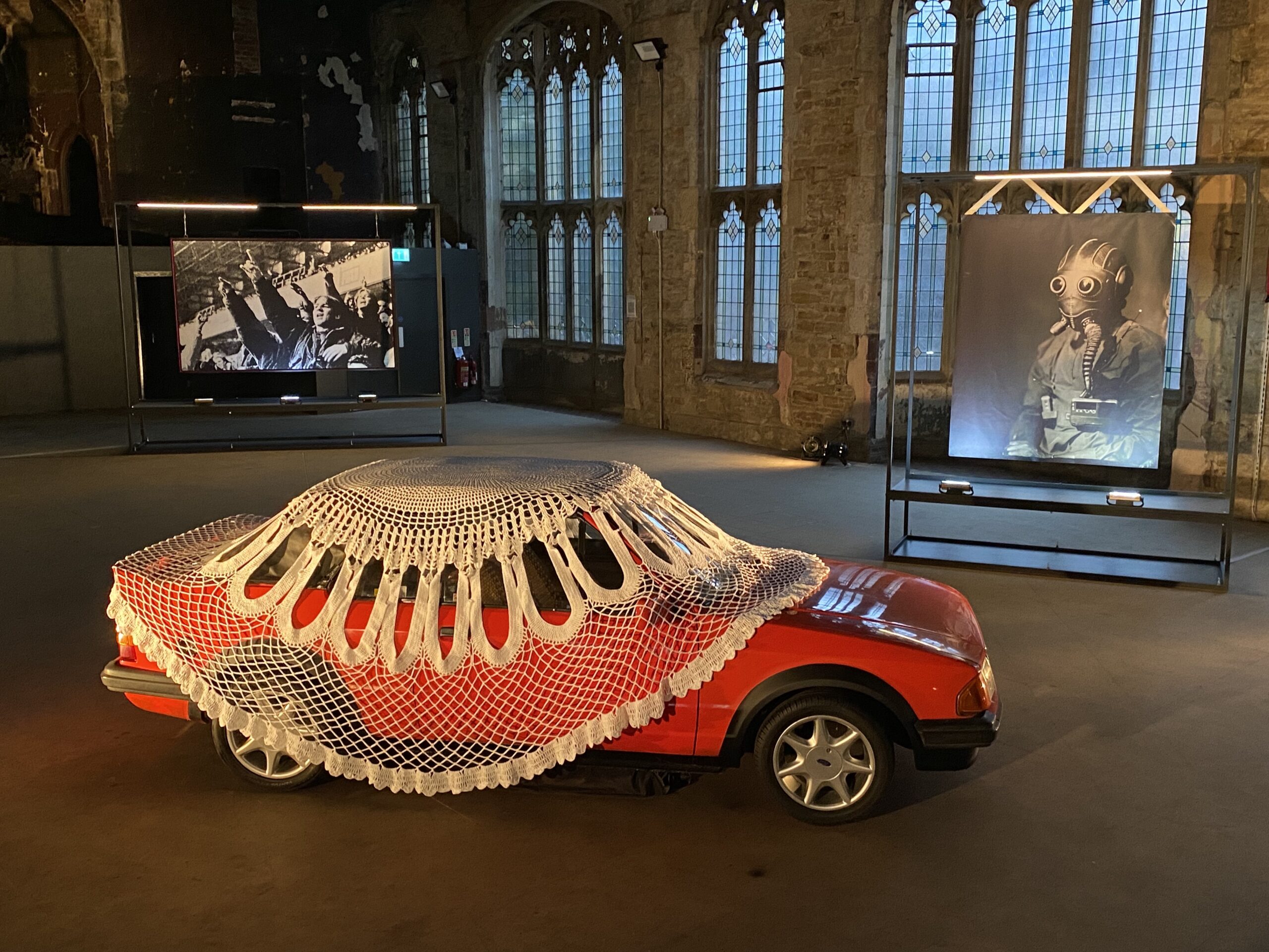 An image of a derelict, formerly grandiose space with a red car covered in a giant doily in the foreground, a photographic portrait of a man in a gask mask in the middle ground, and a tapestry of football fans chanting in the background.