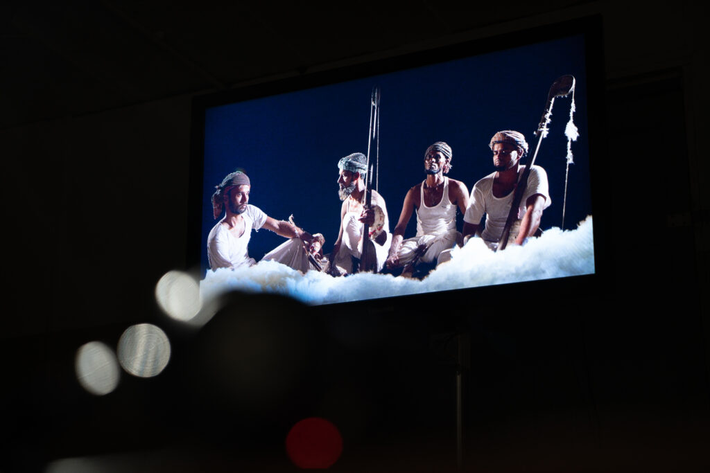 In the middle of a black background the frame of a film can be seen, showing a group of 4 men in the process of calico production.