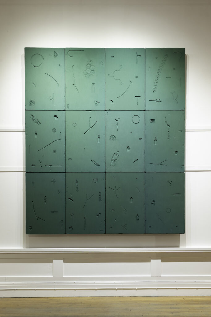 The image shows a rectangle made from 12 pieces of green florist foam attached to a wall. Pressed into each piece of foam are the shapes of various objects found lost in the park.