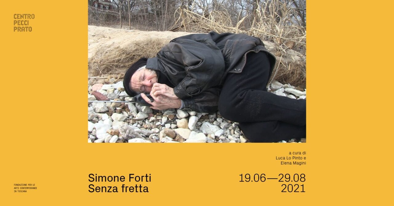 A poster for an exibition with a photograph of the artist curled up on the pebbled ground wearing winter jacket and hat