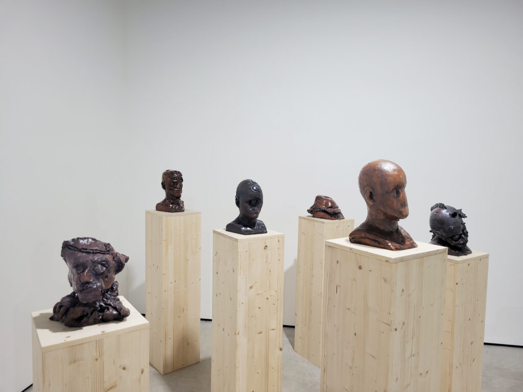 A series of ceramic heads in different stages of addition and deconstruction can be seen place on top of bare wooden plinths, all facing the same direction.