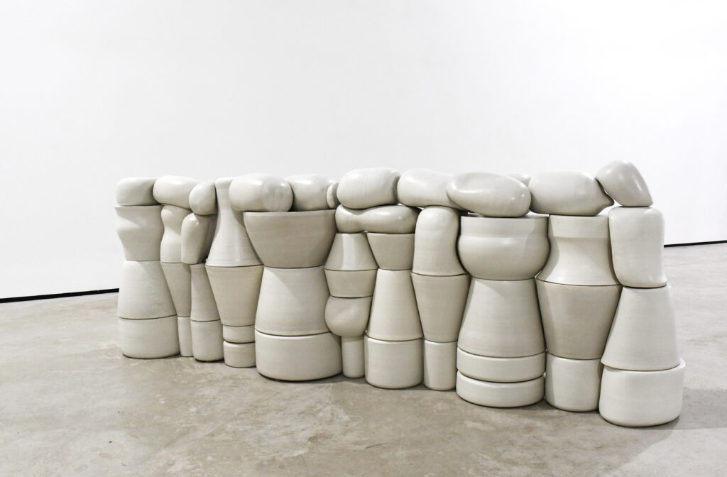 A structure of different ceramic shapes resembling a small wall can be seen, set against the backdrop of a white wall and grey concrete floor.