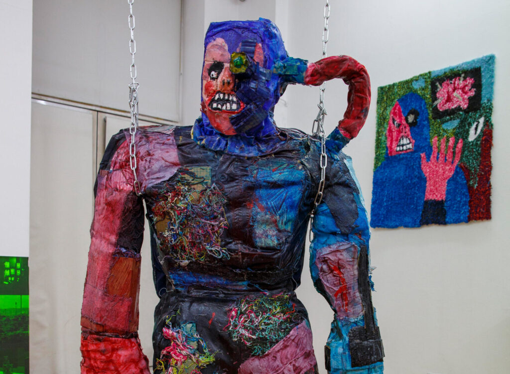 An installation shot from Cyberjunk: Quantum Crash at IMT Gallery: A lifesize figure stands in the foreground of a gallery, the figure is zombie-like and includes robot elements. In the background a rug is mounted on the wall.